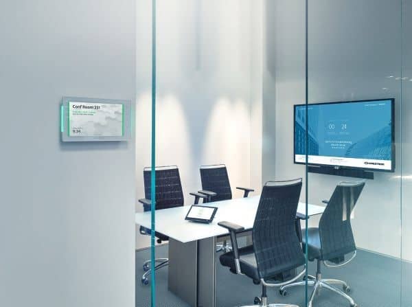 A conference room with glass doors, four office chairs, and a white table with a room scheduling touch panel on the wall.