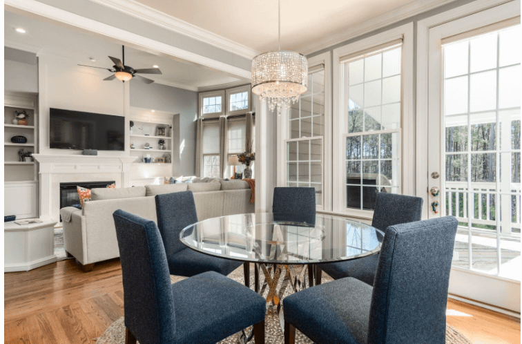 A beautiful well-lit kitchen with a round table, large french doors leading to an outdoor deck and a living room in the background.