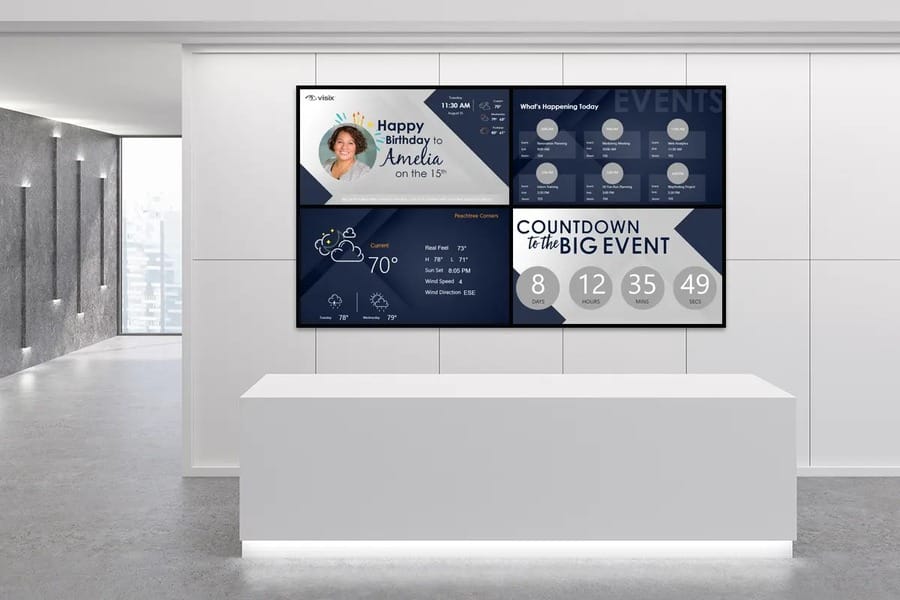 Digital signage displaying someone’s birthday, the weather, an event countdown, and an event calendar above a white desk.